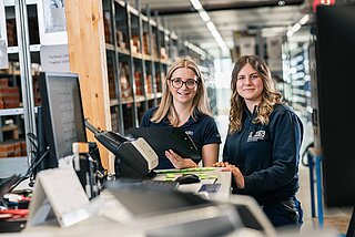 Zwei Mitarbeiterinnen im Lager überprüfen mittels einer Liste, dahinter die Lagerregale