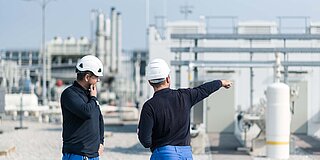 Zwei Montagemitarbeiter mit Sicherheitshelm, einer deutet auf etwas an der Industrieanlage im Hintergrund