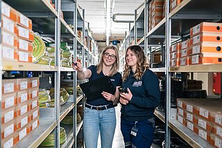 Zwei Lagermitarbeiterinnen deuten auf Ware im Lager zwischen den Regalen