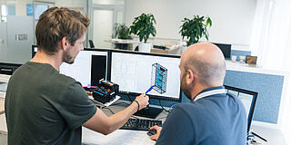 Zwei Automatisierungstechniker im Gespräch deuten auf eine Skizze am Bildschirm, dahinter das Großraumbüro