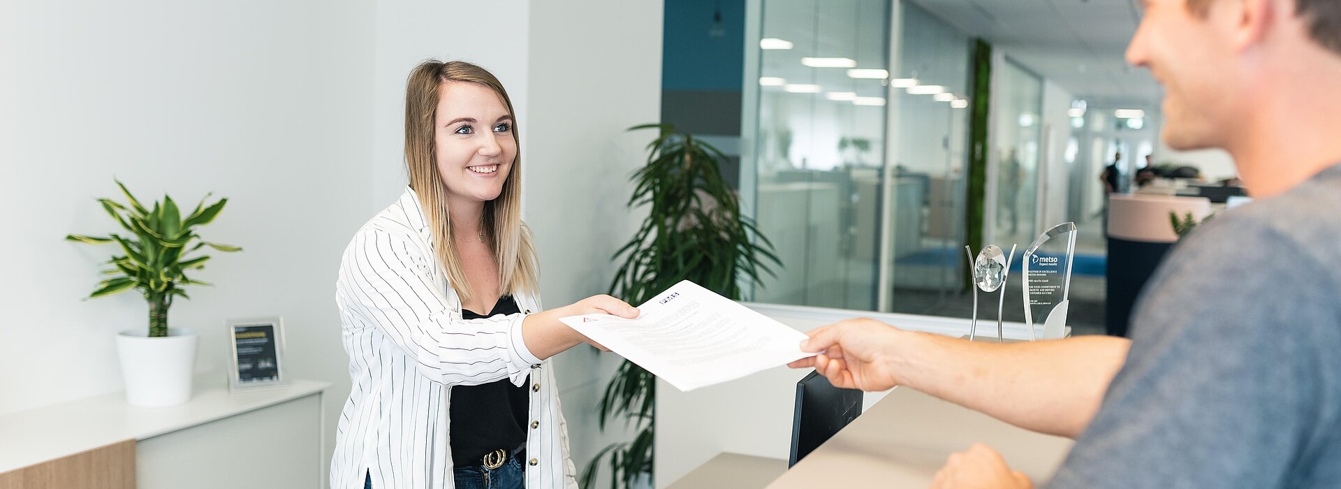 Eine Mitarbeiterin am Empfang übergibt Unterlagen an eine weitere Person vor dem Empfangstisch
