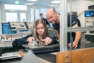Ein Lehrmädchen spannt ein Werkteil in eine Maschine und der Lehrlingsbeauftragte schaut prüfend zu