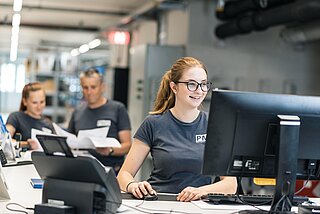 Ene Lagermitarbeiterin arbeitet am PC, dahiner 2 Mitarbeiter, die eine Liste kontrollieren