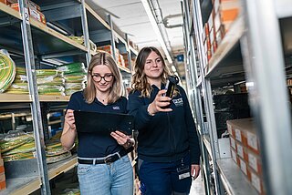 Zwei Lagermitarbeiterinnen scannen Ware im Lager, eine macht sich Notizen in ihren Unterlagen