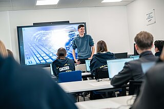 Ein Vortragender unterrichtet die Lehrlinge in der Lehrwerkstätte, die Lehrlinge haben Laptops vor sich am Tisch stehen