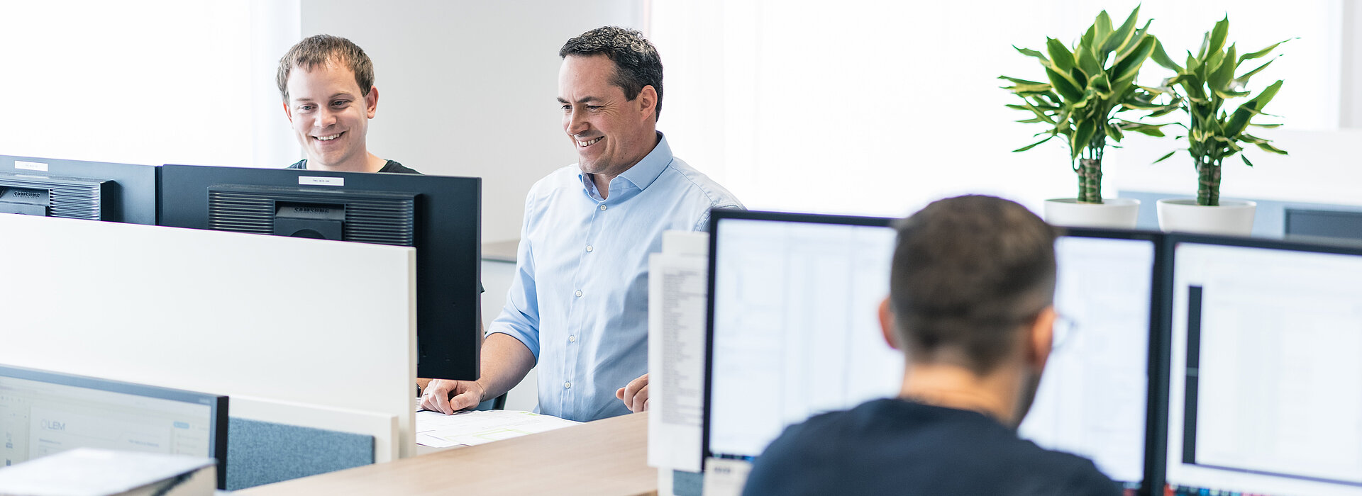 Ein Mitarbeiter beim Arbeiten am PC, dahinter unterhalten sich zwei Kollegen über etwas, dass sie am Bildschirm betrachten